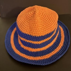 May include: A crochet bucket hat featuring a vibrant orange crown and a wide brim. The design includes alternating horizontal stripes of deep blue and orange yarn throughout the body and the brim, creating a distinct colour-blocked pattern.