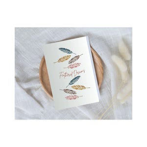 May include: A white card with the text "Feathered Dreams" in red script. The card is decorated with colorful feathers in shades of blue, gold, and pink.
