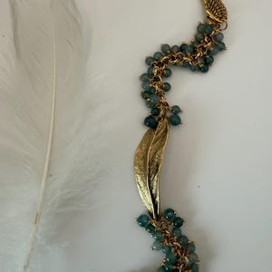 Puede incluir: Un pendiente de oreja dorado con un diseño de ala, conectado a una cadena adornada con pequeñas cuentas de color verde azulado. También se adjunta un dije de hoja dorada. El pendiente y la cadena se colocan sobre un fondo blanco con una pluma blanca.