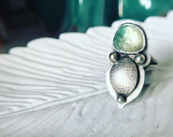 May include: A silver ring featuring a green gemstone and a textured silver design. The ring is displayed on a white surface.