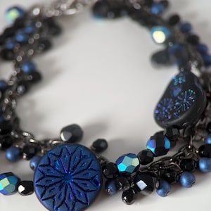 Op de afbeelding: Een donkerblauwe en zwarte kralenarmband met een zilveren kettingsluiting. De armband heeft een grote, ronde, donkerblauwe knoop met een sterrenpatroon, samen met zwarte en blauwe kralen in verschillende maten.