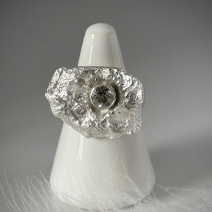 May include: A silver ring with a textured, organic design featuring a clear gemstone in the center. The ring is displayed on a white ceramic ring holder.