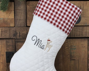 May include: White quilted Christmas stocking with a red and white checkered cuff. The stocking is personalized with the name "Mia" embroidered in gray, along with a small deer illustration. A festive holiday decoration.