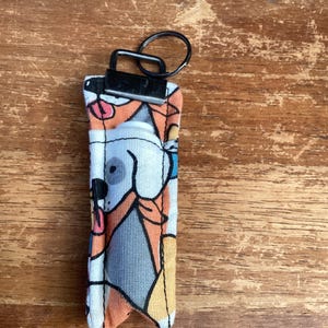 May include: A small, fabric keychain with a colourful cartoon dog print. The keychain has a metal ring and a clip for attaching to a bag or keys.