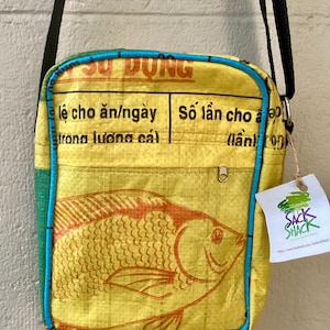 May include: Yellow crossbody bag featuring a fish illustration in orange and brown. The bag has a blue trim and a black strap. Text in a foreign language is printed on the bag. A tag with the text "Sack Shack" is attached.