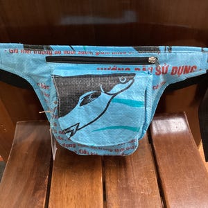 May include: A light blue bum bag made from woven material featuring a black graphic of a fish on the front pocket. The bag has a black zip closure, black adjustable straps with a plastic buckle, and red text printed across the surface.