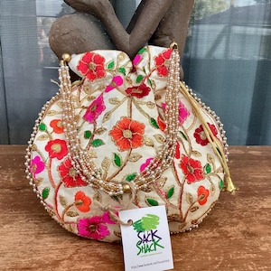 May include: A white drawstring bag with a floral pattern in shades of pink, red, orange, and green. The bag has a gold beaded drawstring and a gold tassel. The bag is sitting on a wooden surface. The bag has a tag attached to it that says "Sack Shack".