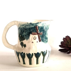 May include: A white ceramic pitcher with a blue and green design featuring a windmill and flowers. The pitcher has a handle and a rounded body.