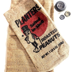May include: A tan burlap sack featuring the Planters logo, a red circle, and the text Salted In-Shell Roasted Peanuts, Net Wt. 1 lb 8 oz. Three small circular metal weights are positioned to the right, labeled 4 oz, 2 oz, and 1 oz.