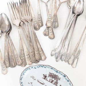 May include: A collection of silver-toned metal forks and spoons with ornate floral handles arranged above an oval ceramic dish. The dish features a blue scalloped rim and brown illustrations of garden items with the text Happiness your Garden filled skies.