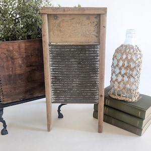 May include: A vintage wooden washboard with a corrugated metal scrubbing surface. The washboard is flanked by a wooden box with greenery and a decorative bottle cover. A stack of green books is also visible.
