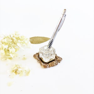 Puede incluir: Un bolígrafo plateado se encuentra en un portaplumas de vidrio transparente con una base de metal dorado. La base tiene un diseño ornamentado.