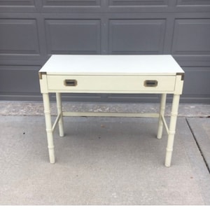 May include: Cream-colored desk with a single drawer and brass hardware. The desk has a rectangular top and bamboo-style legs. The desk is approximately 40 inches wide, 20 inches deep, and 30 inches tall.