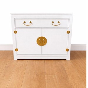 May include: A white cabinet with two doors and a drawer, featuring gold-colored hardware. The cabinet has a rectangular shape with rounded corners and is set against a white wall and a wooden floor.