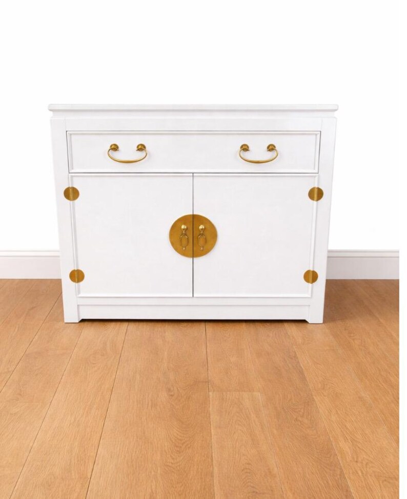 May include: A white cabinet with two doors and a drawer, featuring gold-colored hardware. The cabinet has a rectangular shape with rounded corners and is set against a white wall and a wooden floor.