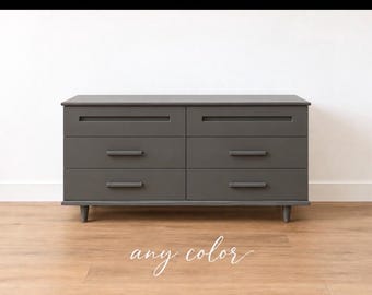 May include: A dark gray six-drawer dresser featuring a rectangular frame, tapered legs, and horizontal bar handles on each drawer. The text "any color" is written in white cursive script below the furniture piece.