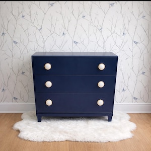 May include: A navy blue three-drawer dresser with white and gold knobs. The dresser sits on a white faux fur rug, with a wallpaper background featuring a tree branch design with small blue birds.
