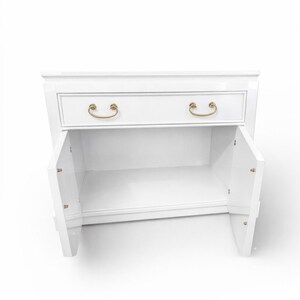May include: A white nightstand with a drawer and two doors. The drawer has two gold-colored handles. The doors are open, revealing the interior. The nightstand is on a white background.