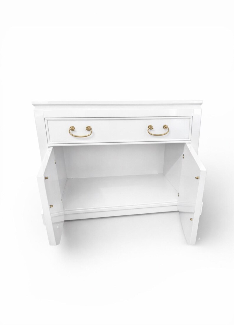 May include: A white nightstand with a drawer and two doors. The drawer has two gold-colored handles. The doors are open, revealing the interior. The nightstand is on a white background.