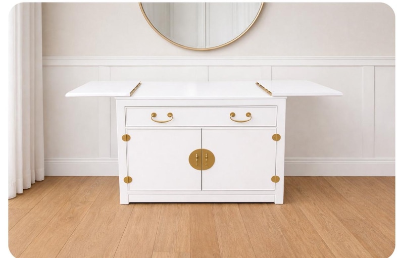 May include: A white cabinet with a fold-out top, two drawers, and two doors. The cabinet has gold-colored hardware, including drawer pulls, door accents, and a central door clasp. The cabinet is set on a wooden floor, with a gold-framed mirror in the background.