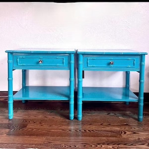 May include: Two matching turquoise nightstands with a bamboo-style design. Each has a single drawer with a silver-colored knob and a lower shelf. The nightstands are set against a white wall and rest on a dark wood floor.