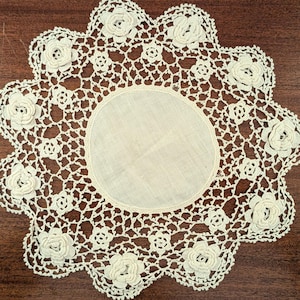 May include: Cream-colored, handmade doily with a floral design. The doily features a central circular fabric piece surrounded by an openwork lace pattern. The edge is adorned with raised, three-dimensional rose motifs, creating a delicate and vintage aesthetic.
