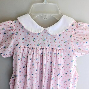 May include: A vintage pink dress with a white Peter Pan collar and short, puffed sleeves. The dress is adorned with a repeating pattern of small blue flowers and red accents. The dress is hanging on a clear plastic hanger.