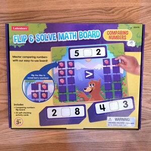 May include: A Lakeshore Flip & Solve Math Board for comparing numbers. The box features a bear illustration and includes a flip board and self-checking activity cards. The board has numbers and berry counters. For ages 3+.