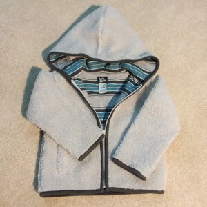 May include: A light blue fleece hooded jacket with a full zipper closure. The jacket has a gray trim and a striped interior lining with blue and teal accents. The brand label is visible on the interior lining.