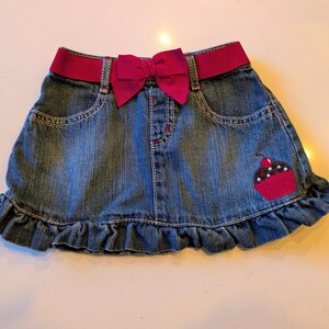 May include: A denim skirt with a dark pink waistband and bow. The skirt has a ruffled hem and a cupcake applique with a cherry on the side. The skirt has two front pockets.