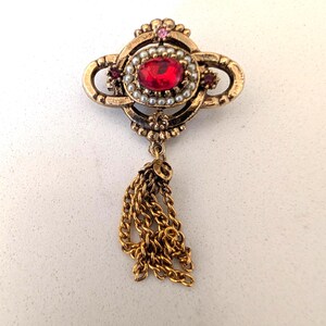 May include: A gold-toned metal brooch featuring an oval-shaped red center stone surrounded by a ring of small white faux pearls. The brooch includes a dangling gold chain tassel and small pink and red accent stones set within the metal frame.