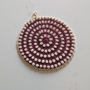 May include: A round, handmade trivet with a spiral design. The trivet features a deep burgundy center with white accents and a beige border. It has a small loop for hanging. The trivet is likely made of yarn or fabric.