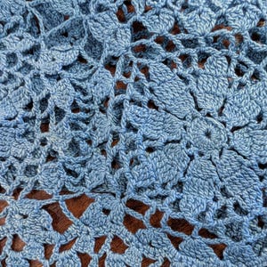 May include: Close-up of a light blue crocheted fabric with a floral pattern. The handmade textile features a delicate openwork design, showcasing the intricate details of the yarn and stitch work. The background is a dark brown color.