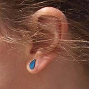 Può includere: Primo piano di un orecchio con un orecchino blu a forma di goccia. L'orecchino è di un blu vibrante ed è inserito nel lobo dell'orecchio. L'orecchio è di carnagione chiara e i capelli sono castano chiaro.