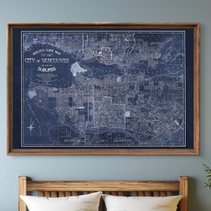 May include: A vintage blue and white map of the City of Vancouver and its suburbs, framed in a dark wood frame. The map is displayed above a wooden bed with gray bedding and white pillows.