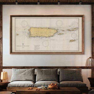 May include: A vintage nautical chart of Puerto Rico and the Virgin Islands, framed in a brown wood frame. The chart is printed on cream-colored paper with brown and black lines and text. The chart is displayed on a wall above a brown couch with gray cushions.