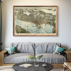 May include: A vintage map of Seattle, Washington, framed in a wooden frame and hanging on a white wall above a gray couch with pillows. The map shows the city's layout and surrounding waterways.