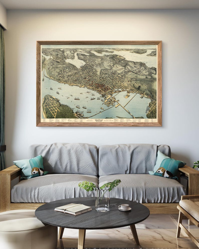 May include: A vintage map of Seattle, Washington, framed in a wooden frame and hanging on a white wall above a gray couch with pillows. The map shows the city's layout and surrounding waterways.
