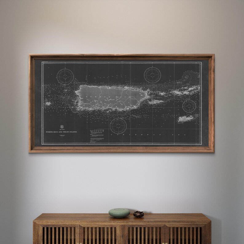 May include: A framed black and white nautical map of Puerto Rico and Virgin Islands. The map is in a dark wood frame and is hanging on a white wall above a wooden cabinet.