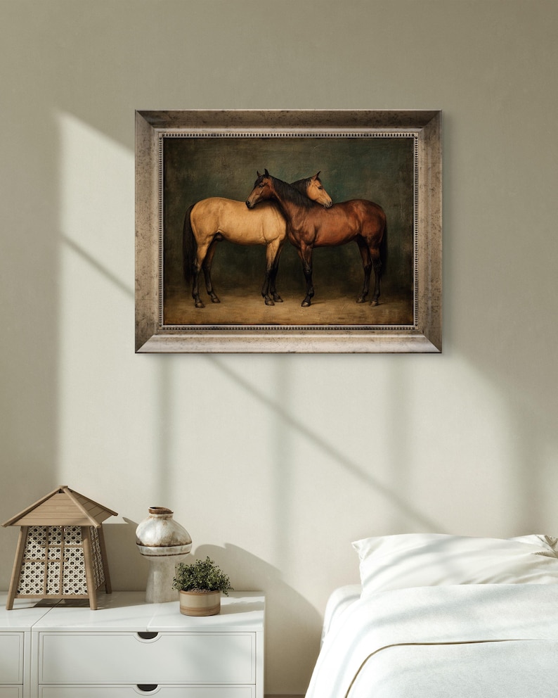 May include: A framed painting depicts two horses, one tan and one brown, standing close together. The horses are set against a dark green background, and the frame is a light, neutral color. The artwork is displayed on a light-colored wall.