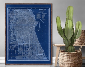 Puede incluir: Un mapa de Chicago, Illinois, de estilo plano vintage, enmarcado en un marco de madera oscura. El mapa está impreso en un fondo azul oscuro con líneas y texto blancos. El mapa está colgado en una pared blanca junto a un cactus en una cesta de mimbre.
