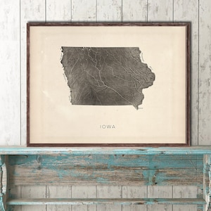 May include: A framed print of a vintage map of Iowa. The map is in black and white and shows the state's rivers and lakes. The word "IOWA" is printed in black at the bottom of the map.