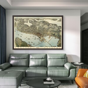 May include: A vintage map of Seattle, Washington, framed in a brown wood frame, hangs on a white wall above a gray sectional sofa with a matching ottoman. A small round black coffee table sits in front of the sofa. A small orange armchair with a yellow and white patterned cushion sits to the right of the sofa.