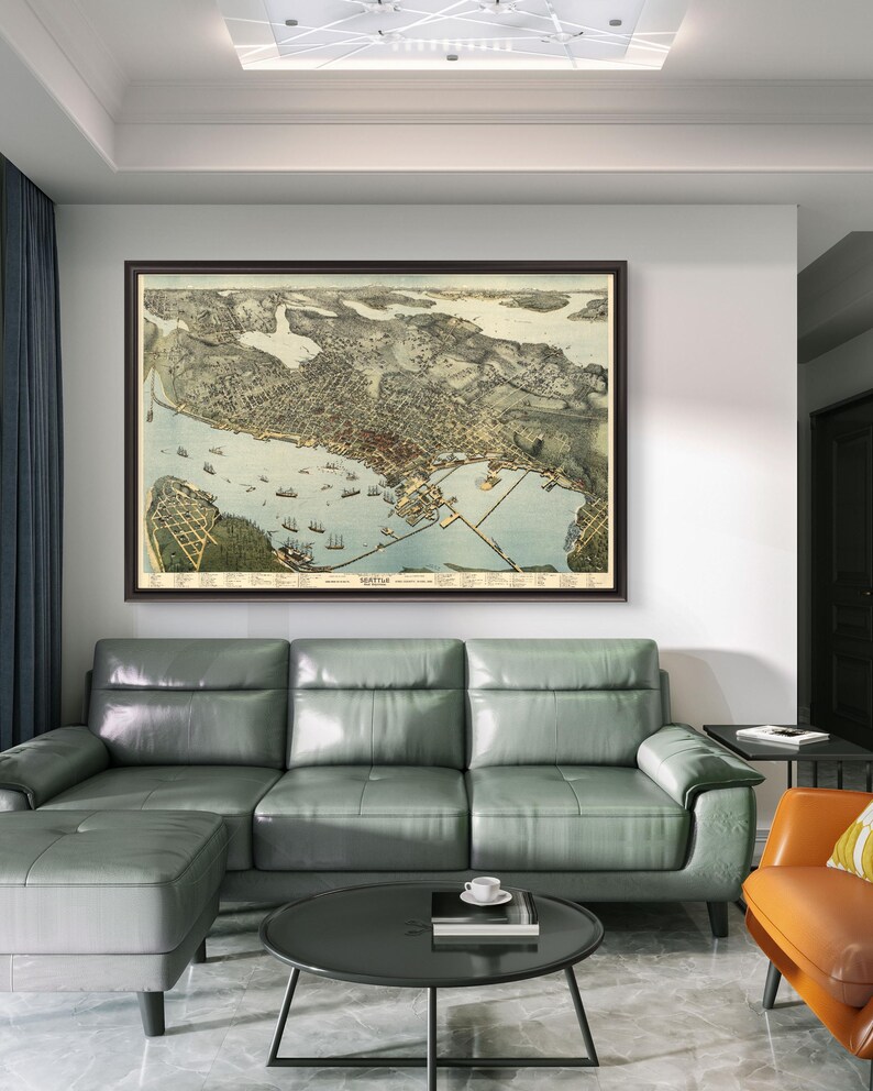 May include: A vintage map of Seattle, Washington, framed in a brown wood frame, hangs on a white wall above a gray sectional sofa with a matching ottoman. A small round black coffee table sits in front of the sofa. A small orange armchair with a yellow and white patterned cushion sits to the right of the sofa.