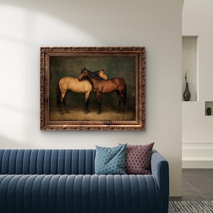 May include: A framed painting depicts two horses, one tan and one brown, standing close together. The artwork has a dark background and is encased in a decorative, ornate brown frame. The painting hangs above a blue sofa with patterned throw pillows.