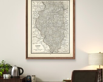 May include: A framed vintage map of Illinois with a brown wooden frame. The map is in black and white and shows the state's roads, cities, and towns. The map is hanging on a white wall above a brown desk with a lamp and a chair.