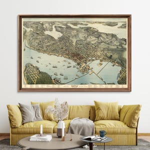 May include: An antique map of Seattle, Washington, showing the city's layout and harbor in detail. The map is framed in a brown wooden frame and hangs on a white wall above a yellow sofa.