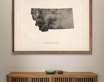 May include: A framed black and white print of the state of Montana with the word "MONTANA" printed below the map. The print is hanging on a light beige wall above a dark brown wooden cabinet with two doors.