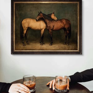 May include: A framed print featuring two horses, one tan and one brown, in a classic painting style. The horses are close together, with one resting its head on the other. The print is displayed above a table with two glasses of a brown beverage.