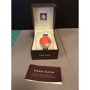 May include: A silver-toned Anne Klein watch with a white leather strap and a vibrant pink and orange face, presented in a branded box. The watch face features a circular design. A warranty card is also included.